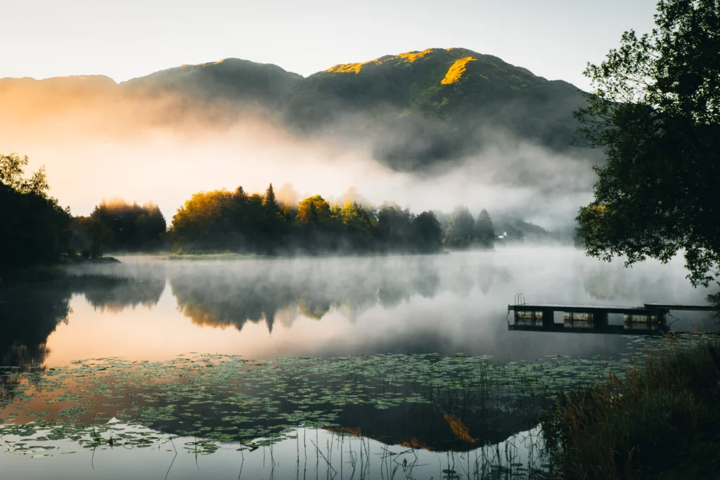 Een mistig meer met zachte reflecties van bomen en bergen, gefotografeerd in diffuus ochtendlicht zonder harde schaduwen - natuur fotografie.